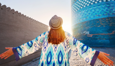 Young woman in ethnic dress with blue ornament and hat spread hand infront of blue Kalta minor minaret in Itchan Kala ancient city of Khiva in Uzbekistan. Credit: Shutterstock